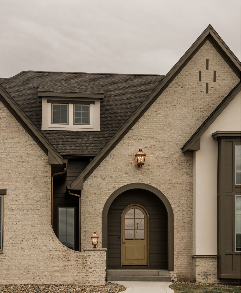 Front view of a house with a brick facade, arched entryway, wooden door, and exterior lights under a cloudy sky.