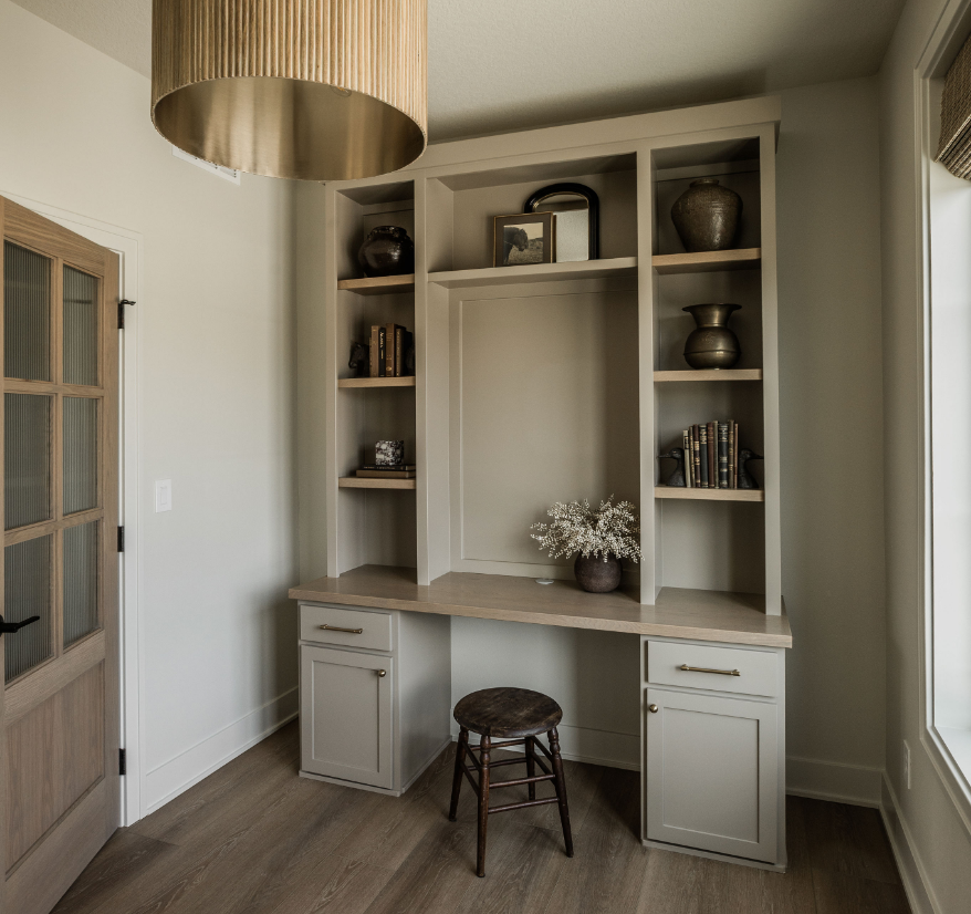 A built-in desk with cabinetry and open shelves is set against a white wall in a room with wood flooring. The shelves contain decorative vases, books, and framed photos. A small stool is in front of the desk, and a window with blinds is to the right.