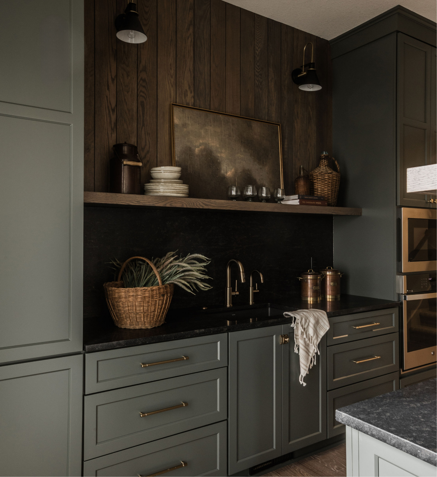 Kitchen with dark green cabinets, a black countertop, and a wooden backsplash with wall-mounted black and gold lights. Decor includes stacked white bowls, a framed artwork, a wicker basket with green plants, glassware, and kitchen utensils.