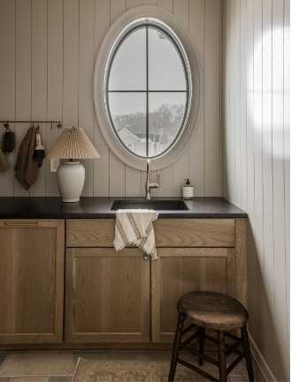 Kitchen with wooden cabinets, a black countertop, a white oval window above the sink, a beige lamp, a dish towel, and a small stool.