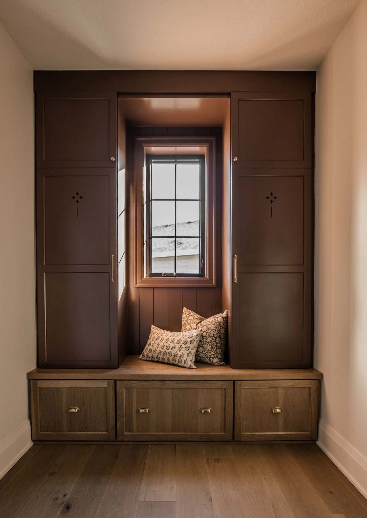 Built-in wooden window seat with cushions and a window with a view of rooftops, framed by dark wooden cabinetry in a cozy interior space.
