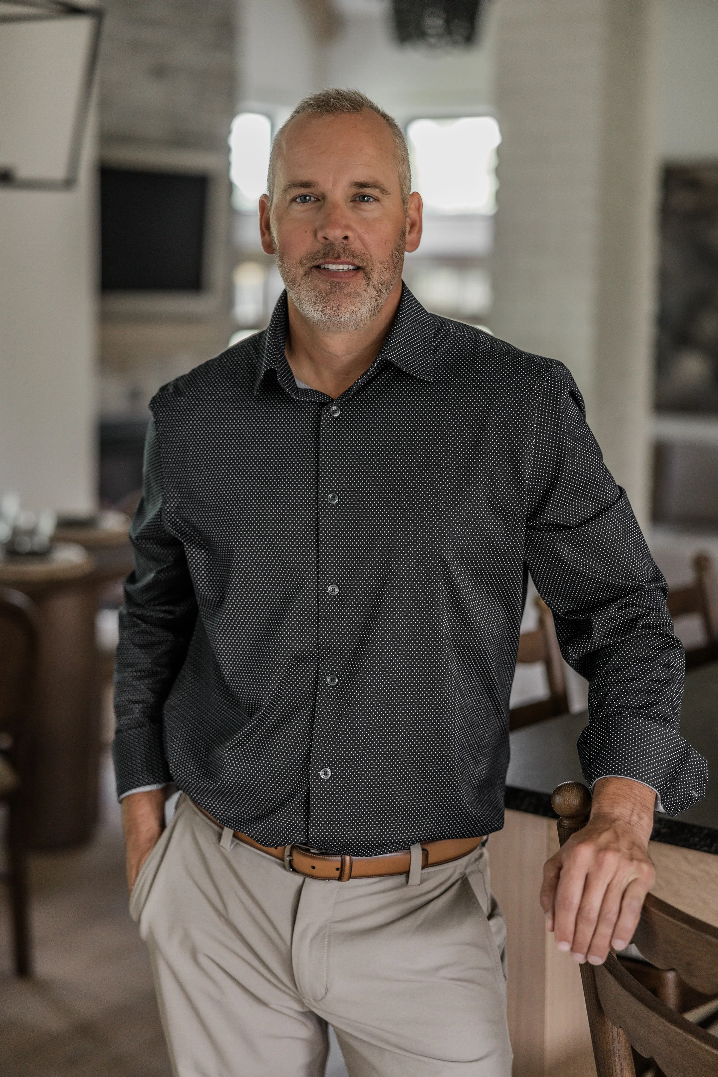 A middle-aged man with a gray beard and short hair, wearing a black patterned button-down shirt and beige pants, standing in a modern indoor space, resting one hand on the back of a wooden chair.