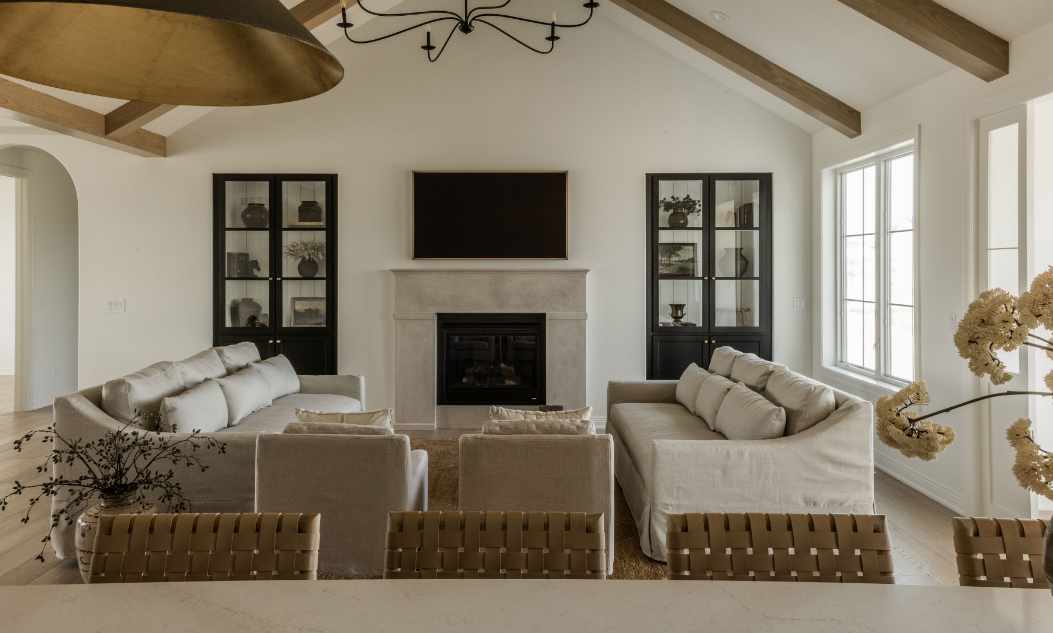 Living room with two beige sofas facing a fireplace and a mounted flat-screen TV, black display cabinets on either side, large windows, and ceiling beams.