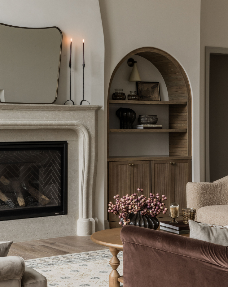 Living room with a beige fireplace, a mirror above the mantel, and two tall black candlesticks. To the right of the fireplace is an arched wooden shelving unit with decor items. In front, there is a round wooden coffee table with a black vase holding pink flowers, a book, and a decorative glass. Sofas and armchairs are partially visible.