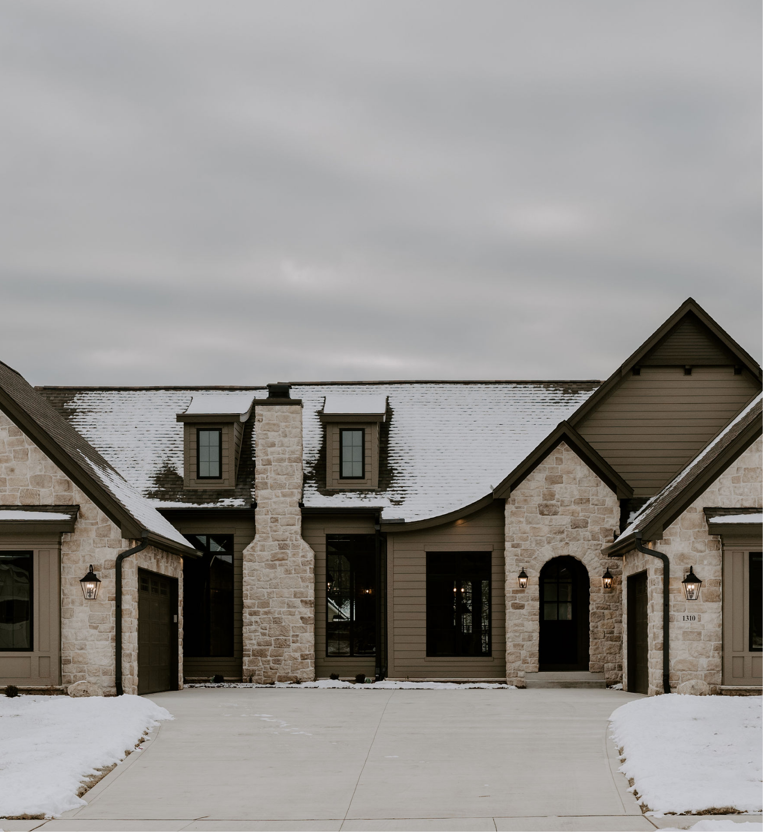 Front view of a house with stone and siding exterior, snow on the roof and ground, with two garage doors and an entrance with a door and windows, outdoor lanterns, and a paved driveway.