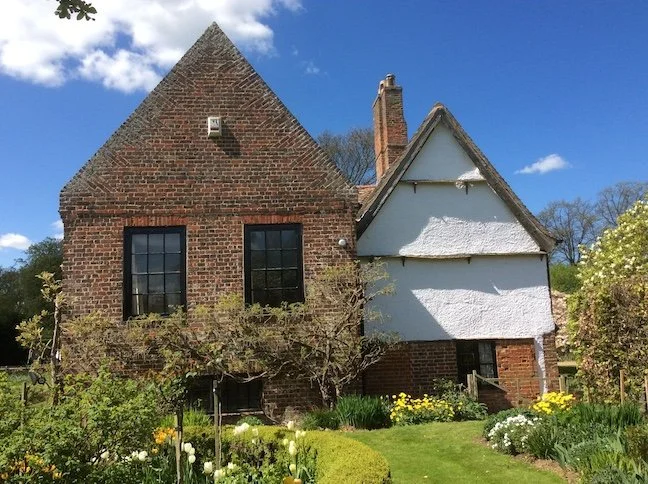 A two-story brick house with a steep, triangular roof on the left, and an attached white stucco section on the right, surrounded by a lush green garden with blooming flowers and trees, under a blue sky with scattered clouds.