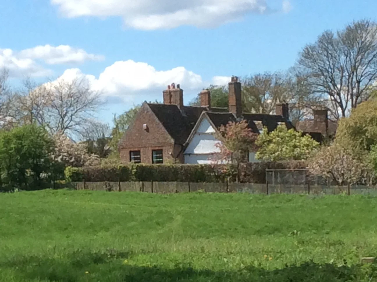 A house with a brick exterior and multiple chimneys, surrounded by trees and a green lawn, under a blue sky with white clouds.