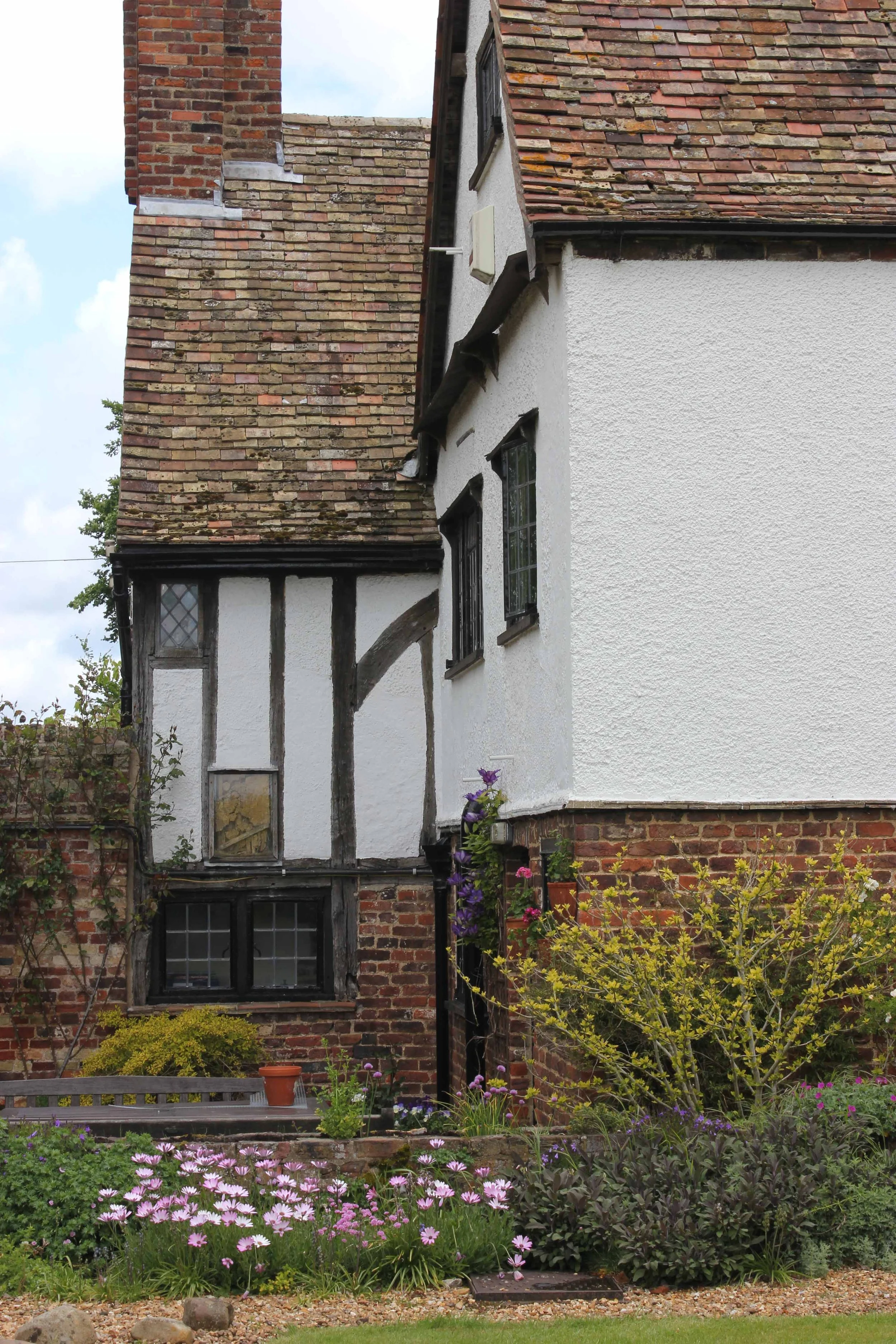 A garden with a variety of colorful flowers including pink and purple, in front of a house with white and brick exterior walls and a red-tiled roof. The house has multiple windows and a small bench with a flowerpot on it, surrounded by trees and plan