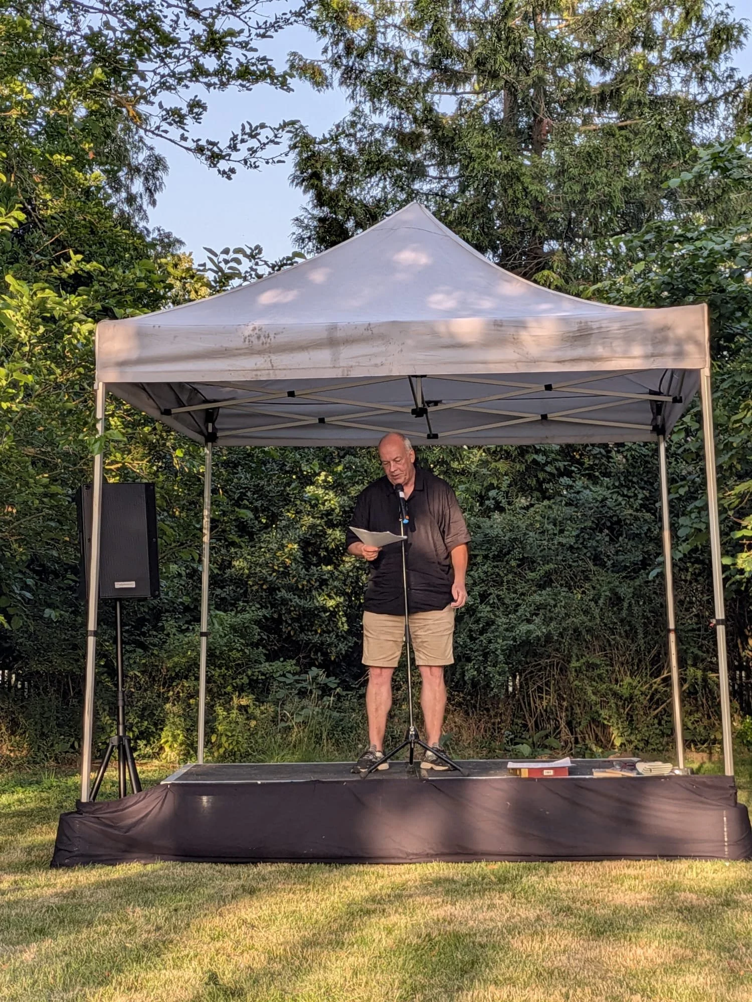 A middle-aged man wearing a black jacket and khaki shorts stands on an outdoor stage under a white canopy, reading from a piece of paper into a microphone, with trees and greenery in the background during daytime.