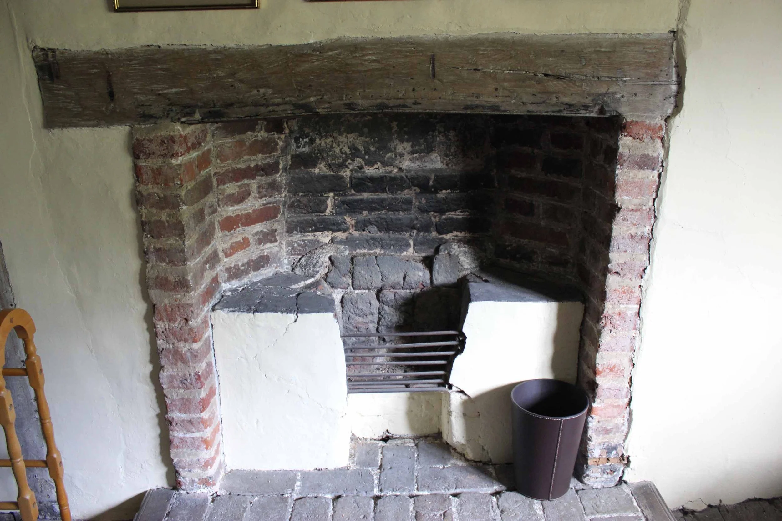 An old brick fireplace with a wooden mantel, inside there are metal fireplace tools, and a black trash can on the right side.