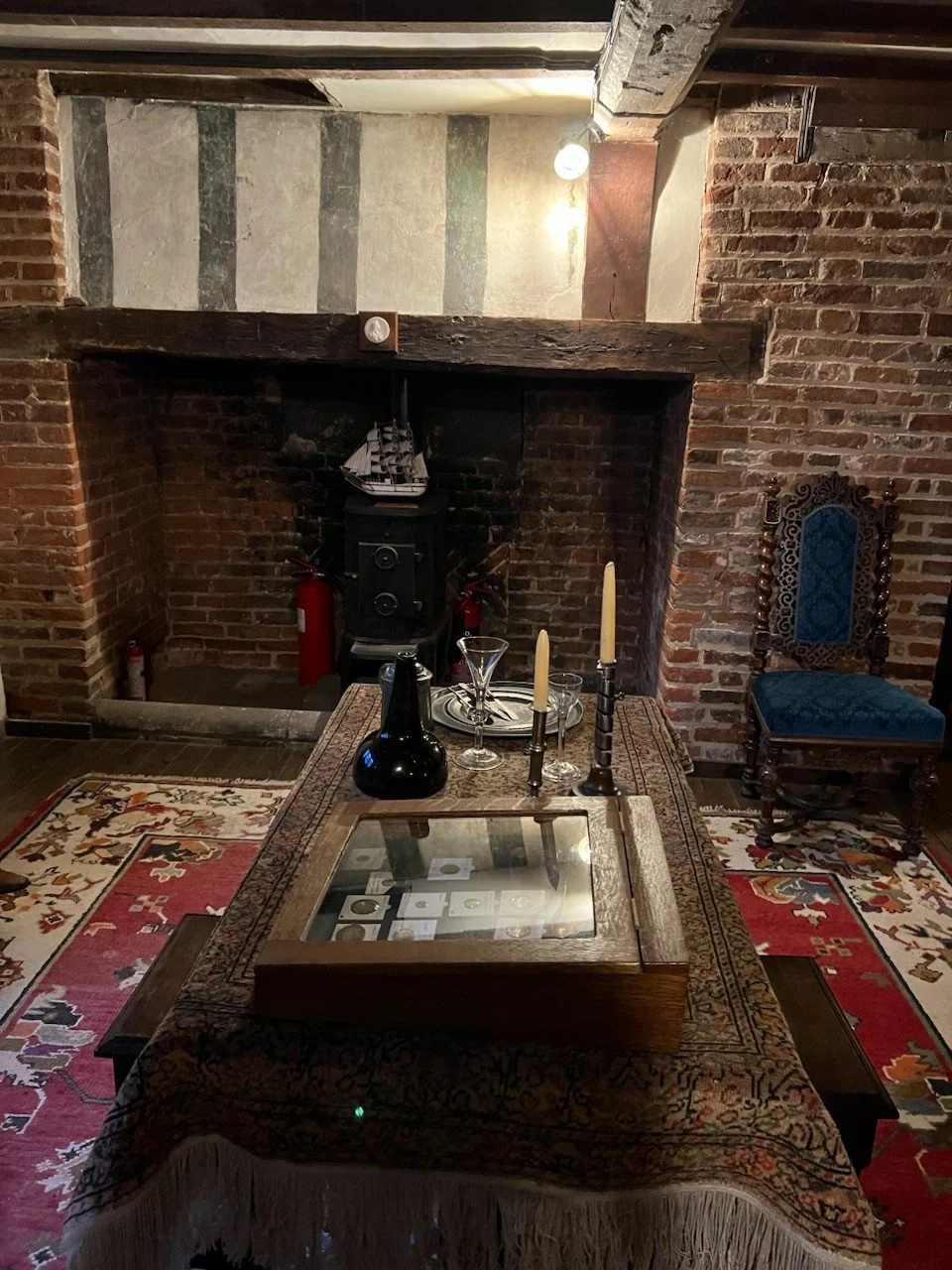 A vintage-style dining room with a brick fireplace, wooden beams, and an ornate blue upholstered chair. A large table with an intricate patterned tablecloth is set with a glass display case, candlesticks, and glassware. The room has a cozy, rustic at