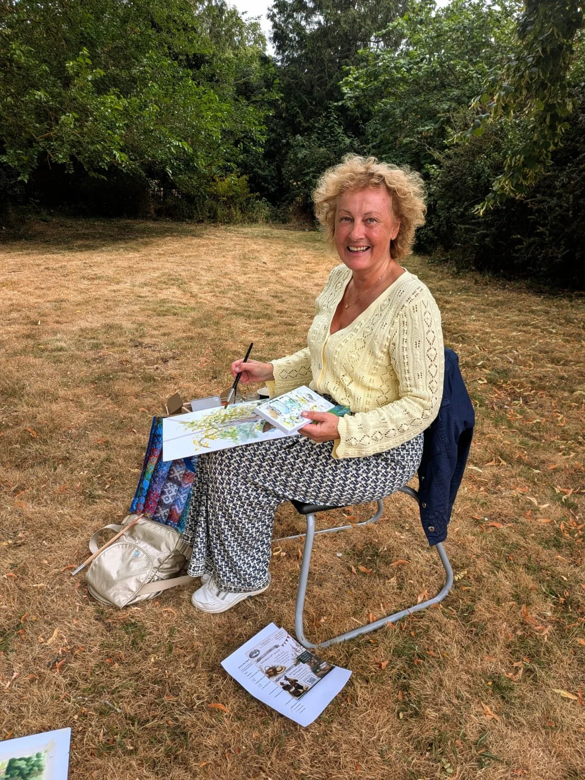 Smiling woman sitting outdoors in a grassy area with trees, painting or drawing on a sketchpad, with art supplies and papers around her.