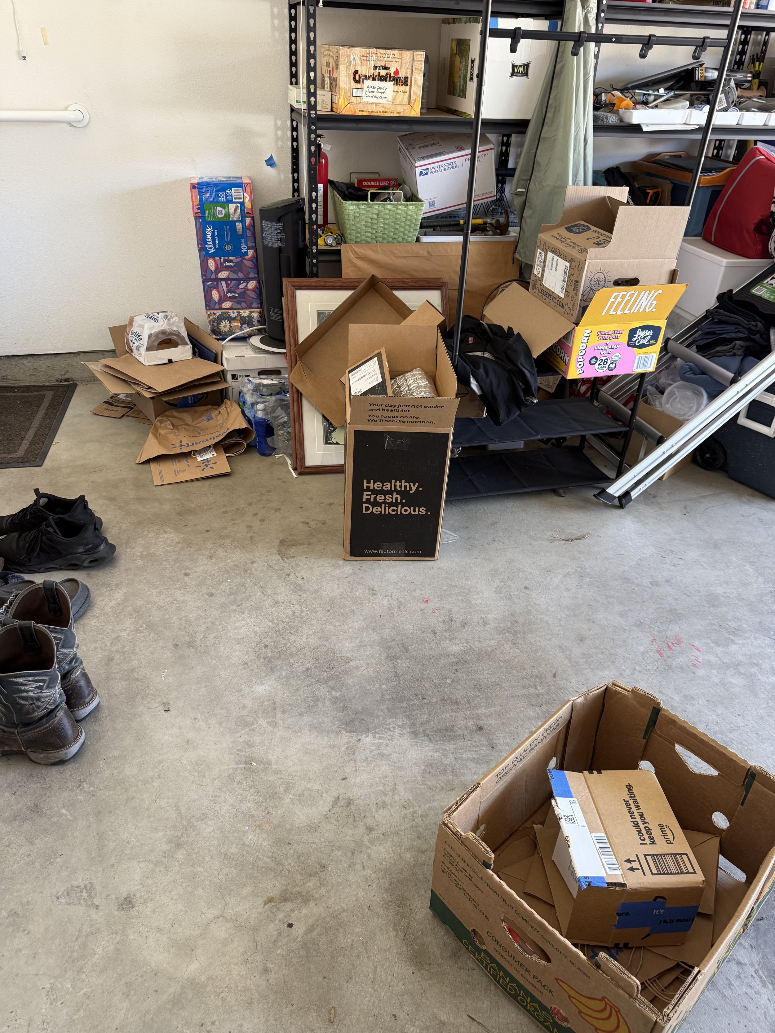 Garage with scattered items including shoe pair, cardboard boxes, picture frames, shelving unit with stored items, and a sign reading 'Healthy. Fresh. Delicious.'