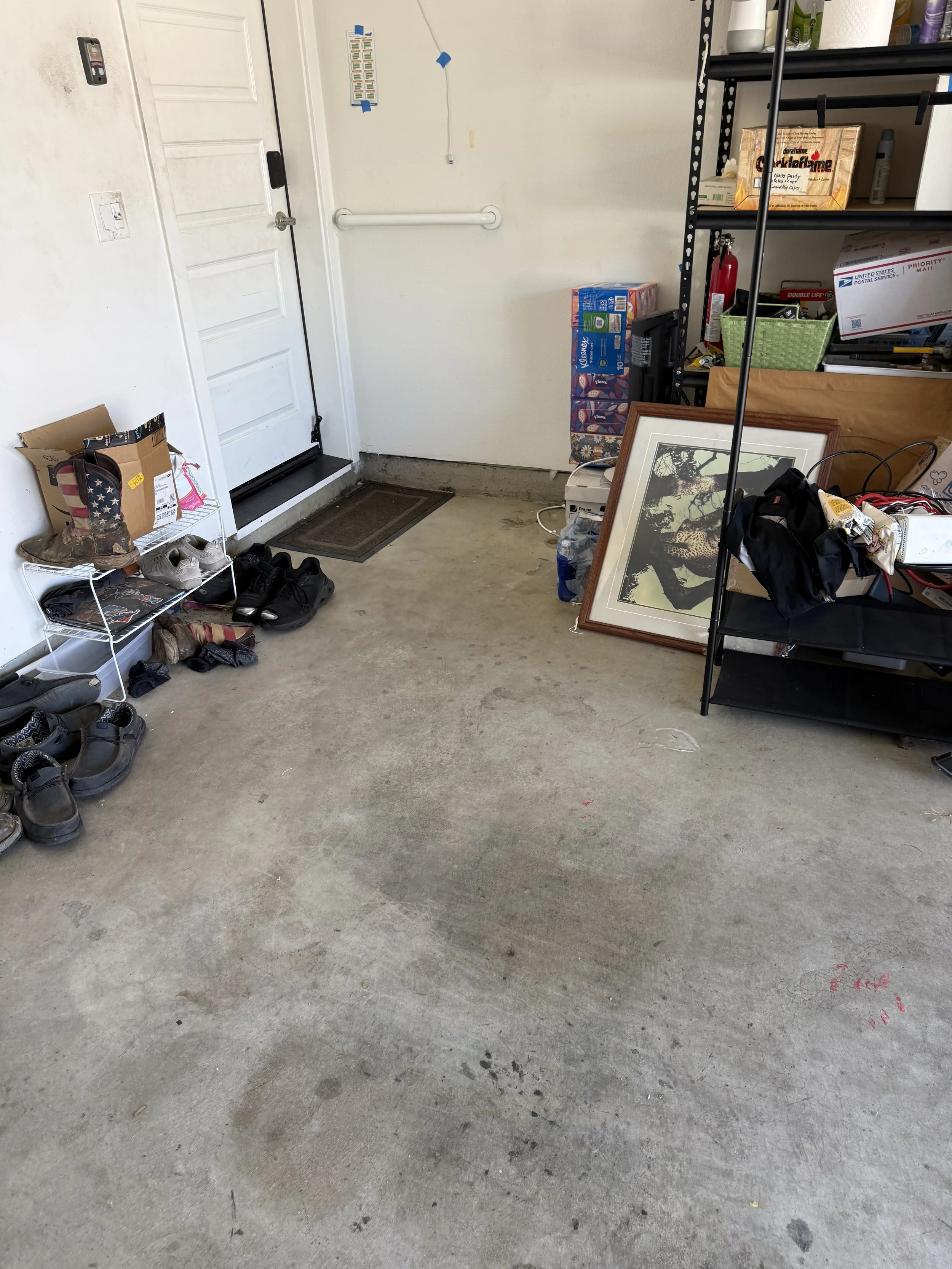 A garage with shoes on a small white wire rack, a framed picture leaning against a shelf, various boxes and supplies on metal shelves, a fire extinguisher, and red marks or stains on the concrete floor.