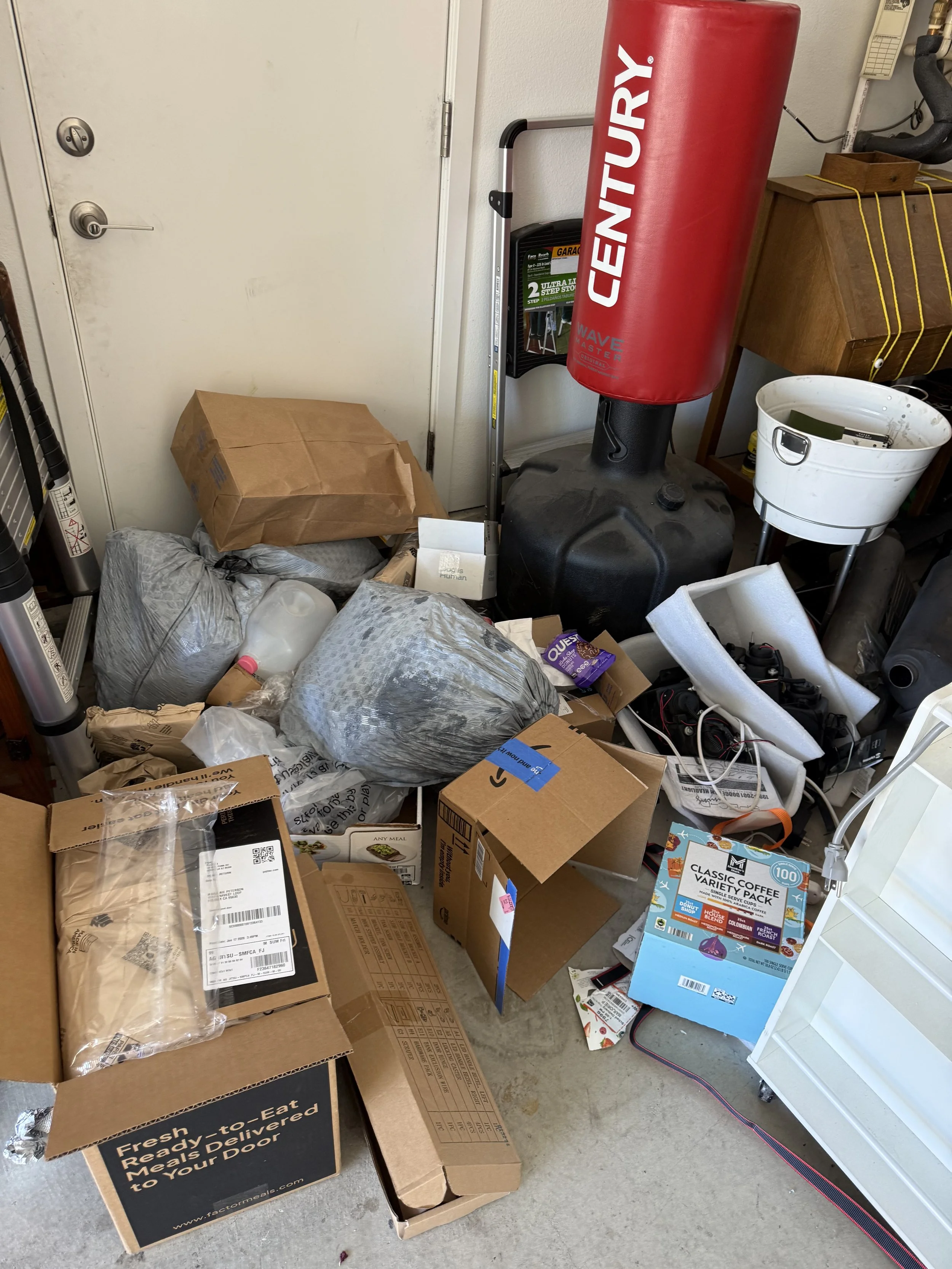 A cluttered garage with cardboard boxes, bags of trash, a punching bag, a step stool, a white bucket, and various miscellaneous items.