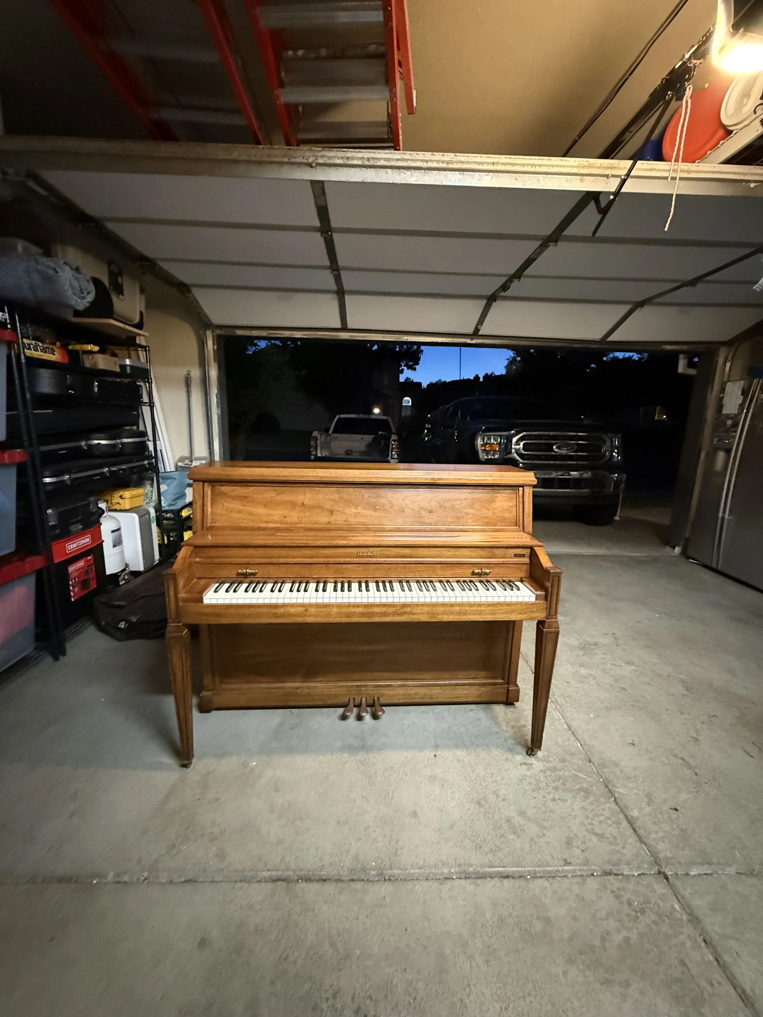 A wooden upright piano inside a garage with a concrete floor and various tools and equipment on shelves to the left, automotive vehicles visible outside through the open garage door, and a ladder hanging from the ceiling.