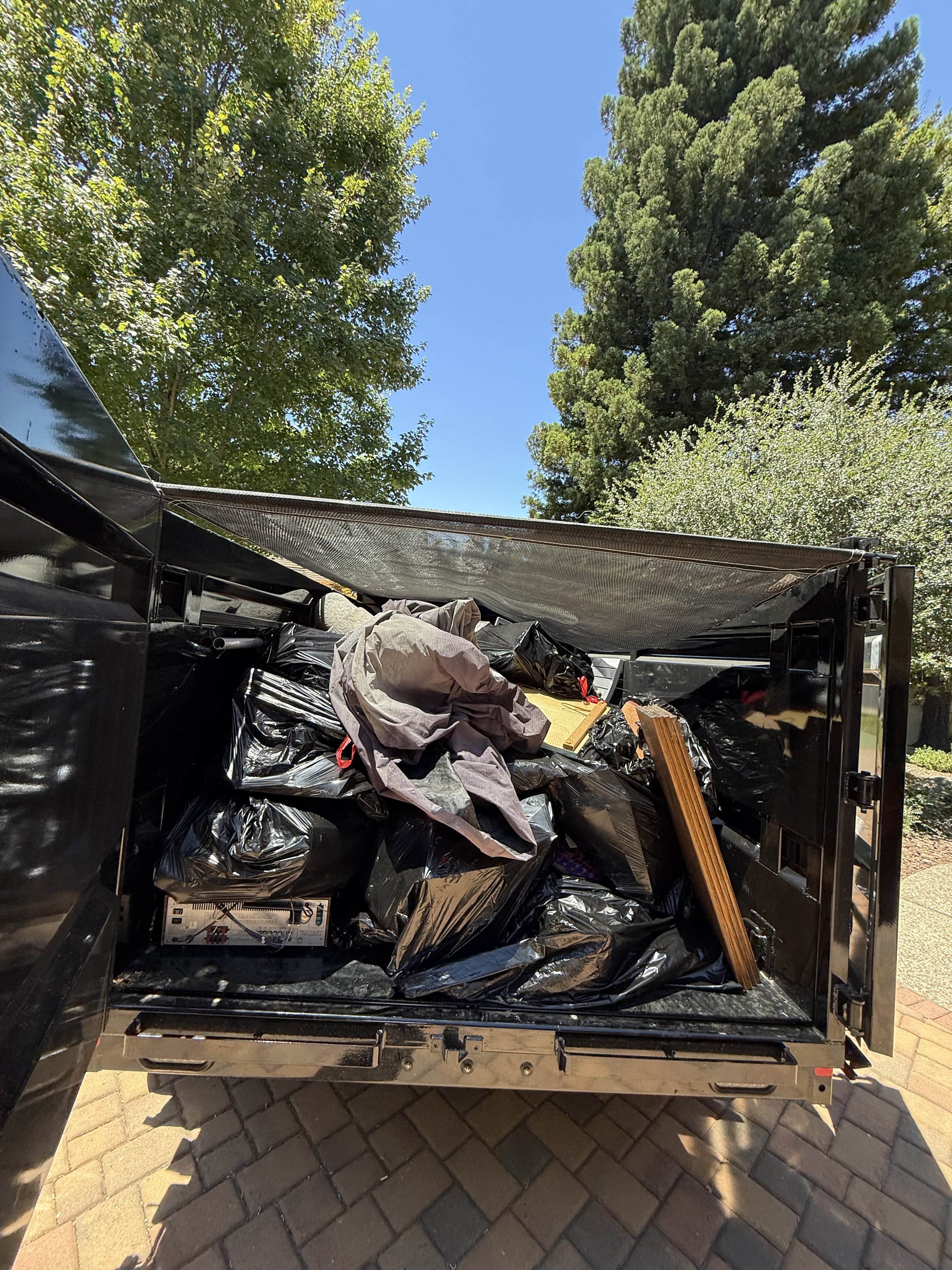 Open truck bed filled with trash and debris, with trees and a blue sky in the background.