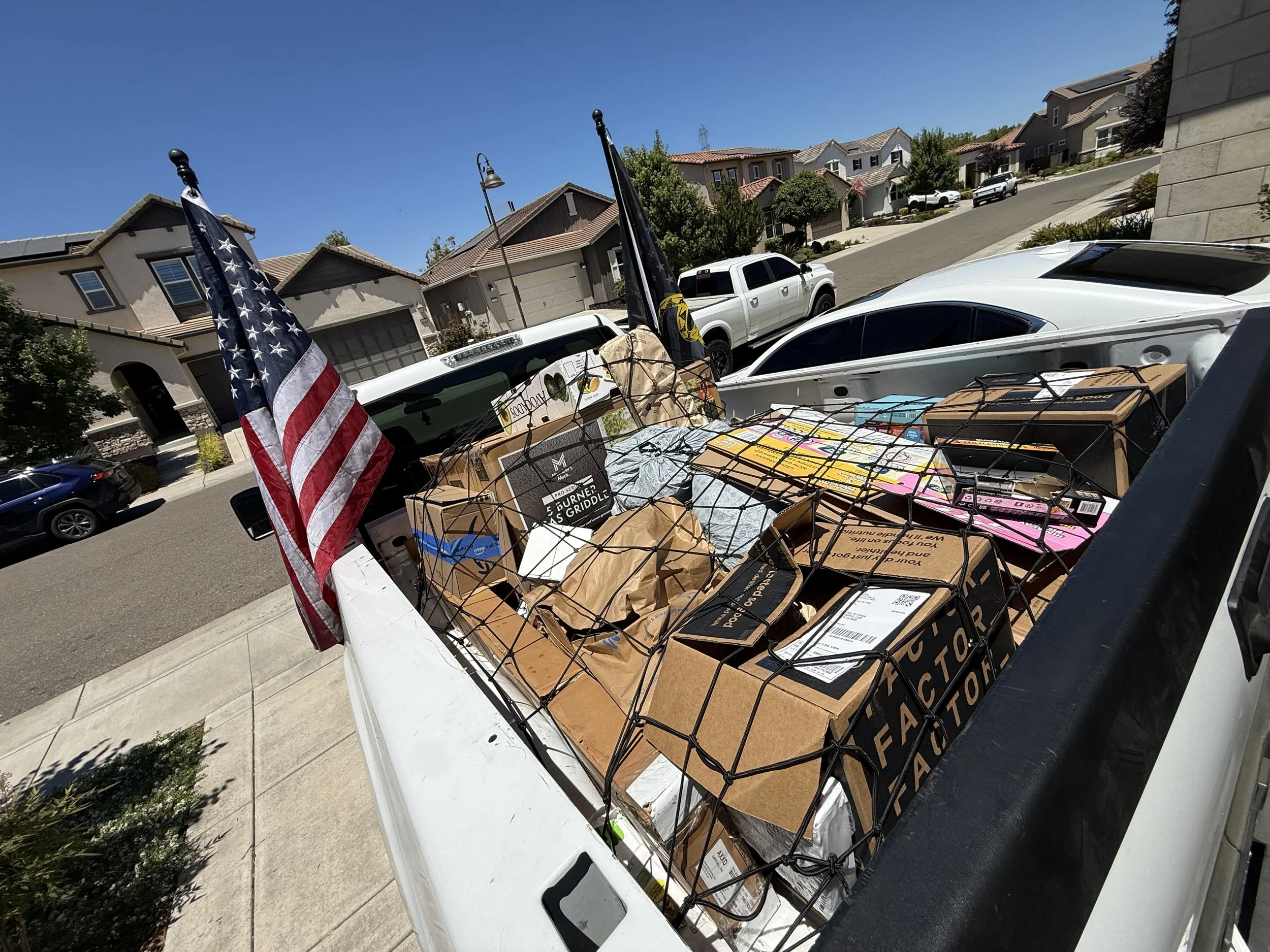 A pickup truck parked on the street loaded with boxes and various items, including American flags and a metal grid, during daytime in a suburban neighborhood.