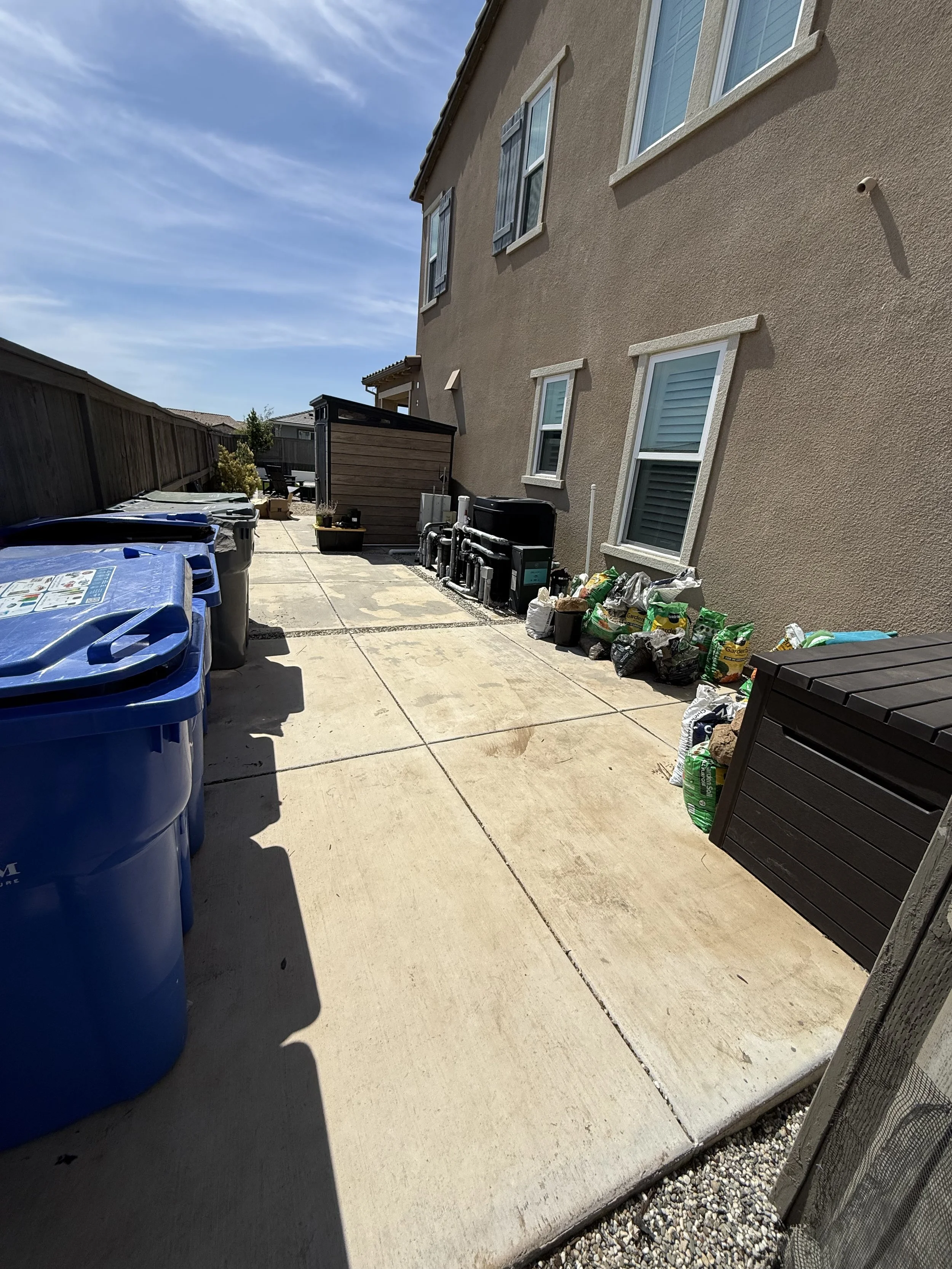 Backyard patio with blue trash and recycling bins, a black storage shed, potted plants, and miscellaneous gardening supplies along the house wall under a clear blue sky.