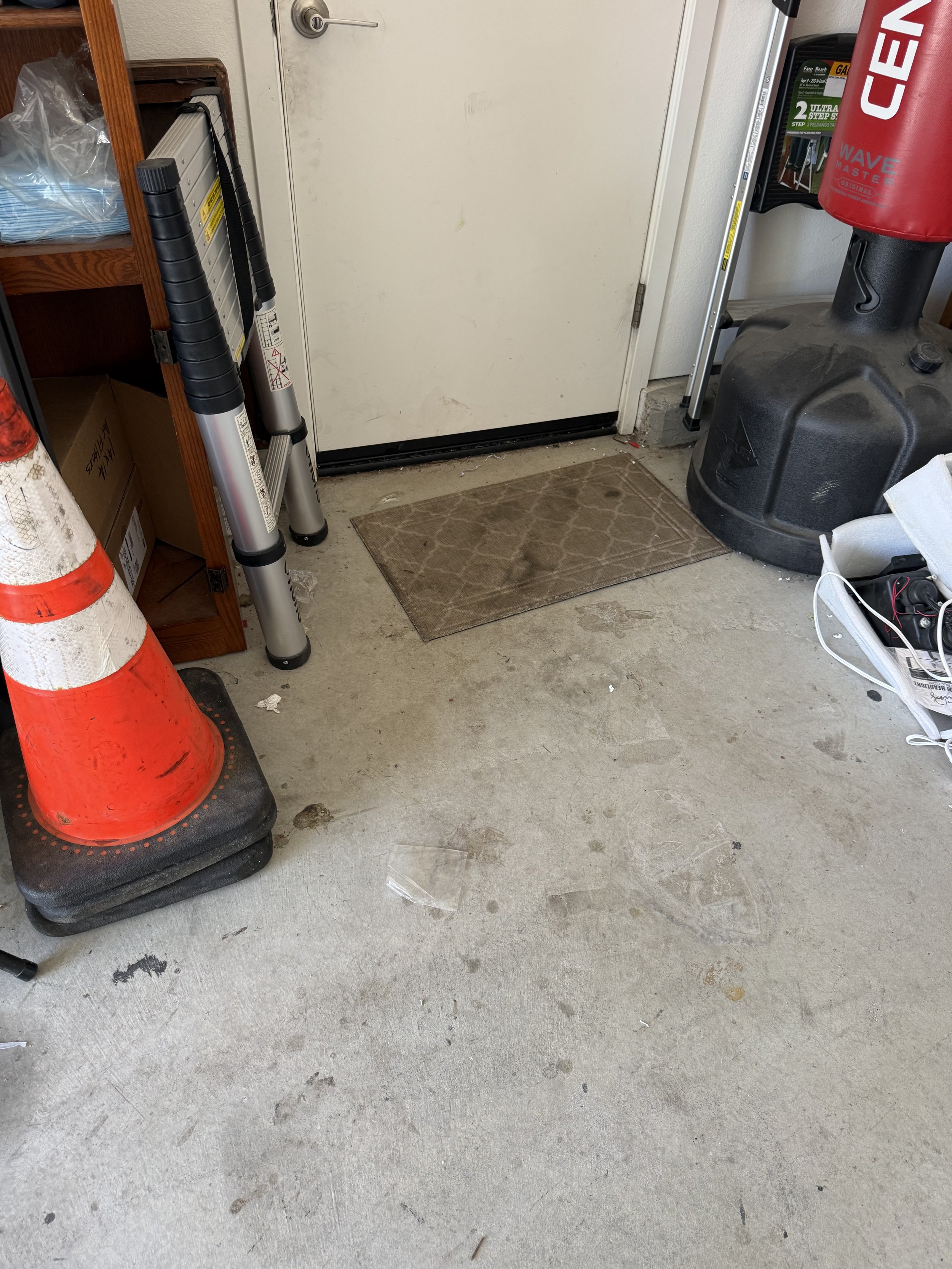 Garage or storage area with orange safety cones, a pair of step ladders, a small rug in front of a plain white door, a cardboard box, a black plastic container, and a white basket with cords, amidst scuff marks and debris on the concrete floor.