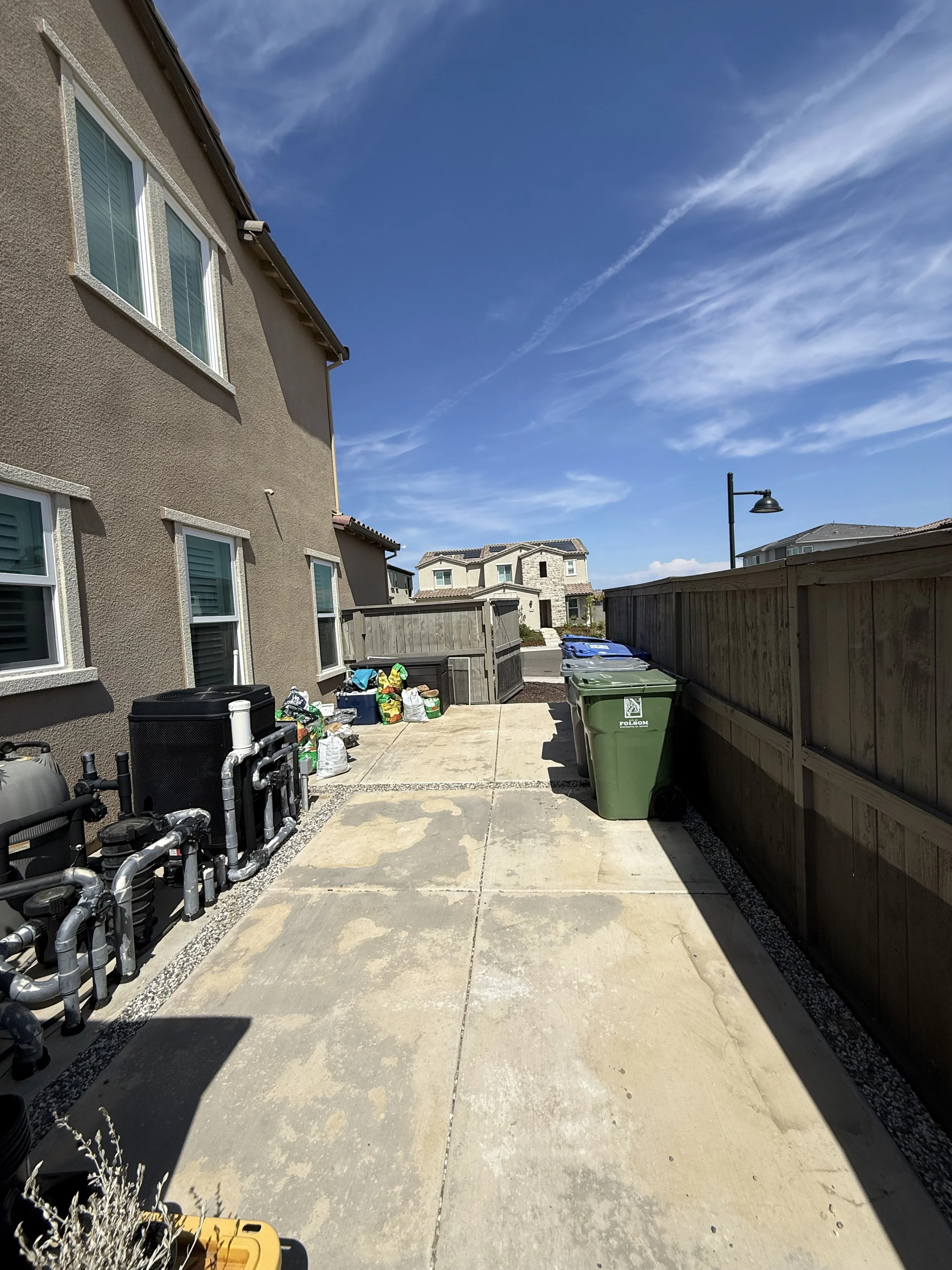 View of a backyard with a concrete patio, green trash bins, a wooden fence, neighboring houses, and a partly cloudy sky.