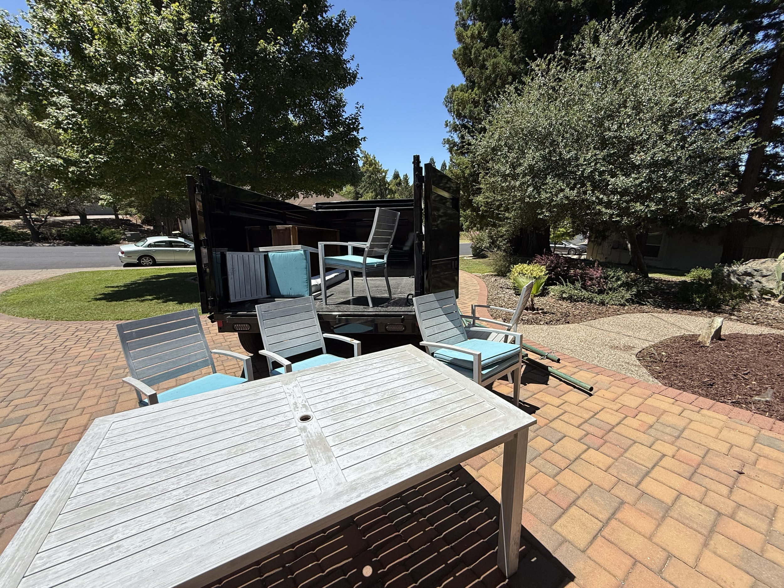 A yard with a patio, multiple chairs, a white table, a truck with stacked furniture, green trees, a blue sky, and a parked car in the background.