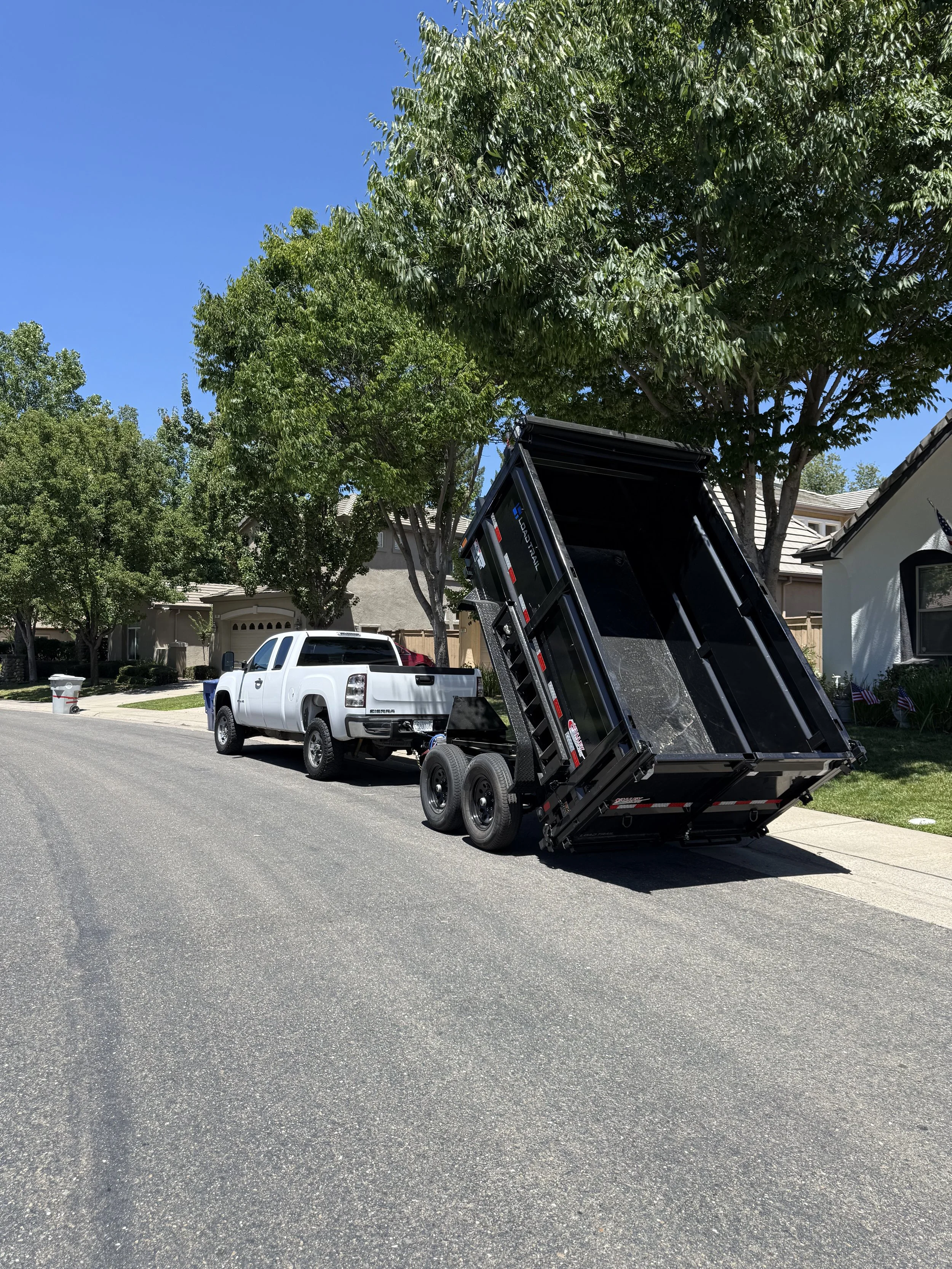 A white pickup truck with a black dump trailer raised, parked on a suburban street next to houses and trees under a blue sky.