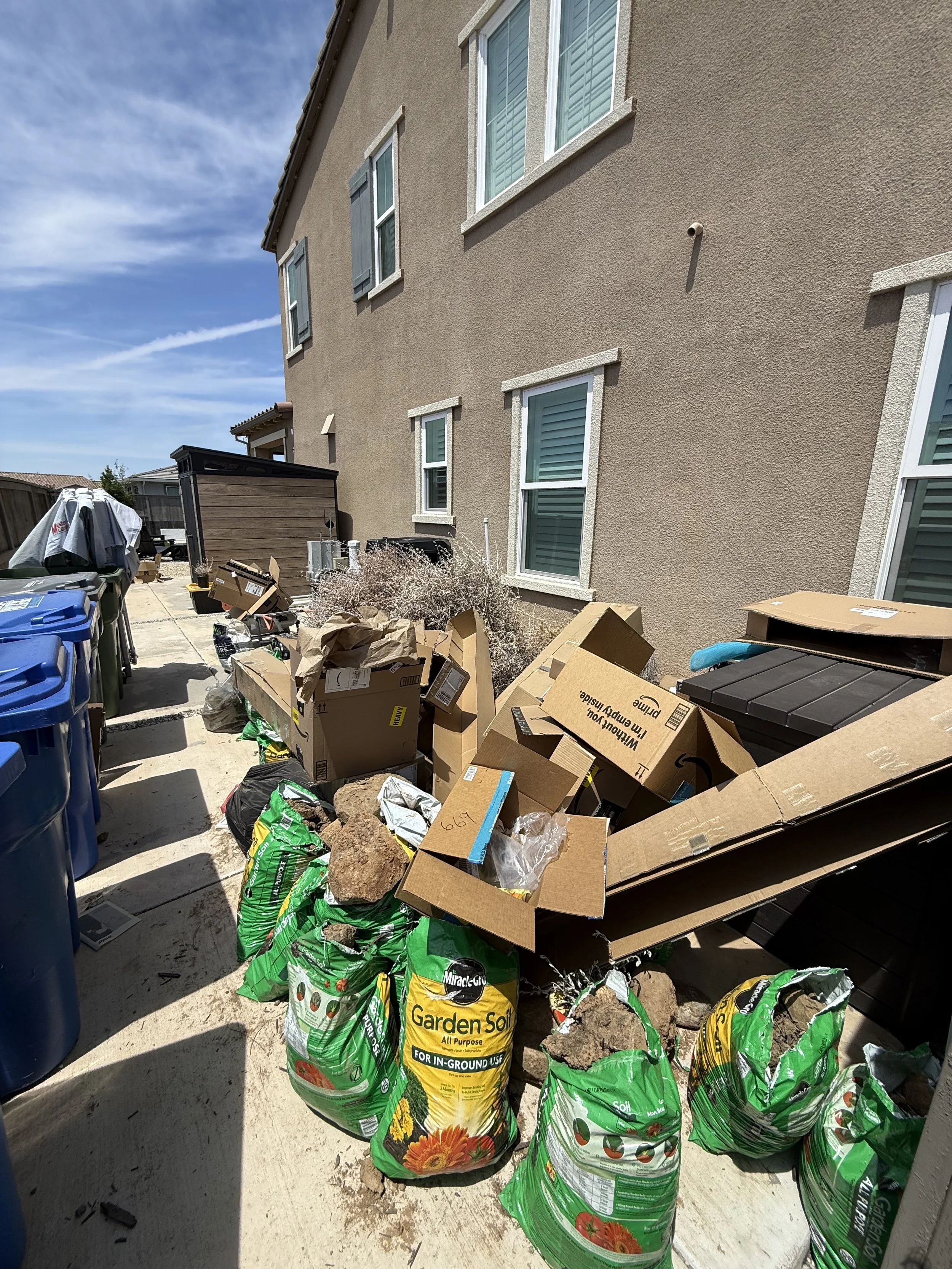 Backyard with house wall, windows, and piles of gardening supplies including soil bags, rocks, and cardboard.
