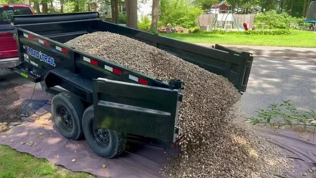 Black trailer filled with gravel dumping onto ground, on a driveway in a backyard with trees and a swing set.