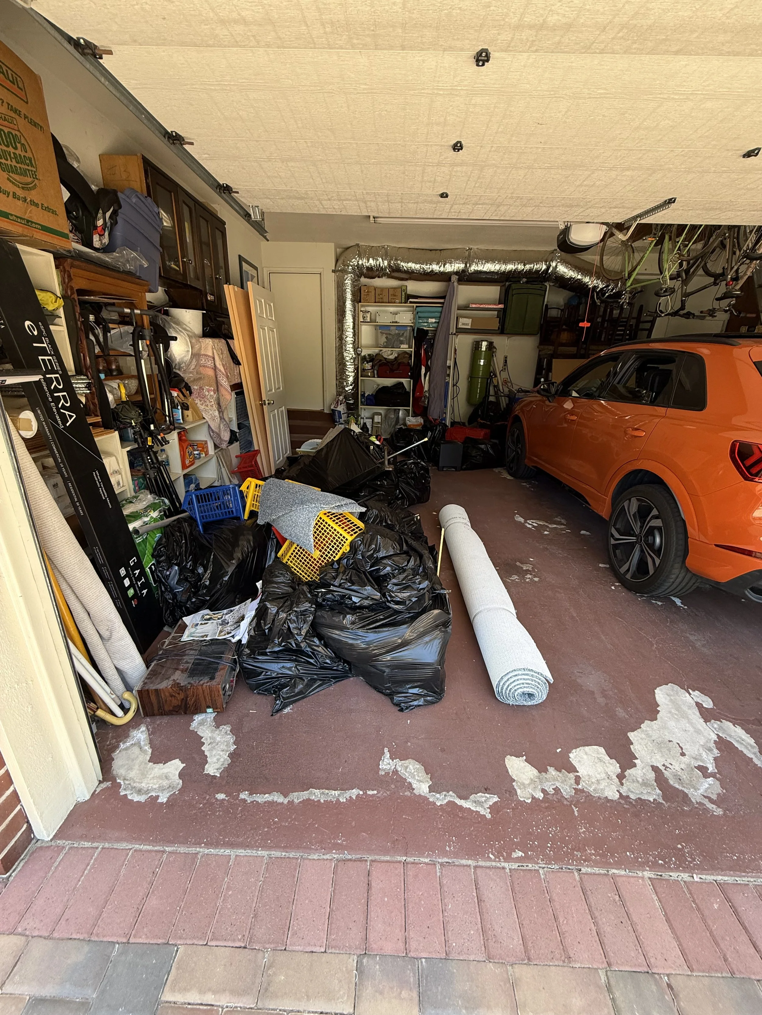 This is an image of a cluttered garage with a bright orange car parked inside. The garage floor is partially painted red, with patches of paint chipping. There are various items scattered on the floor, including black trash bags, a roll of white foam, yellow and blue plastic baskets, and small furniture pieces. The background shows shelves with boxes, a green oxygen tank, and other storage items. The ceiling has several black hooks, and the garage walls are filled with miscellaneous items and equipment.