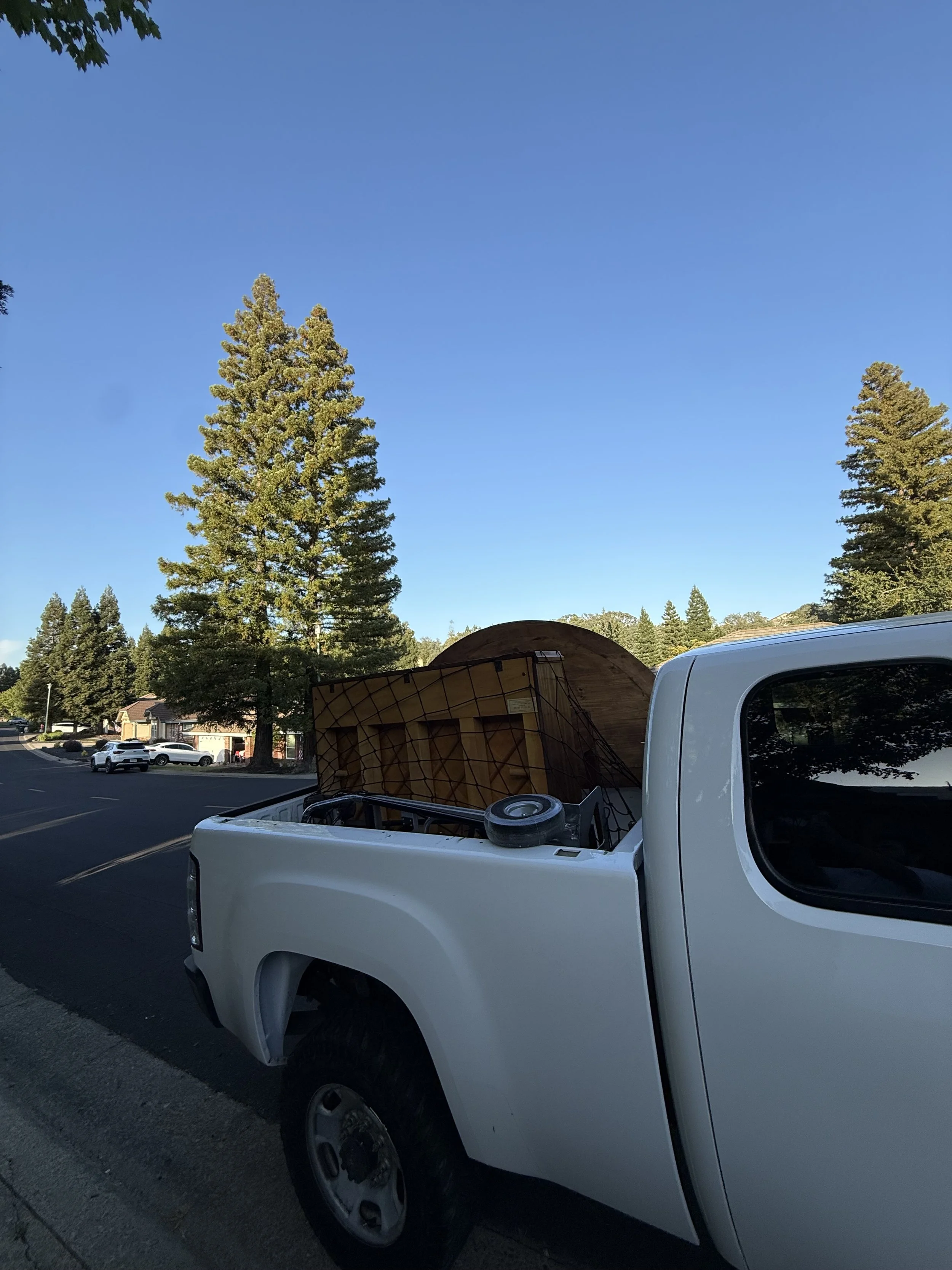 A white pickup truck parked on a street with a tree and a house in the background. The truck's bed has some objects, including a wooden structure and a small wheel.