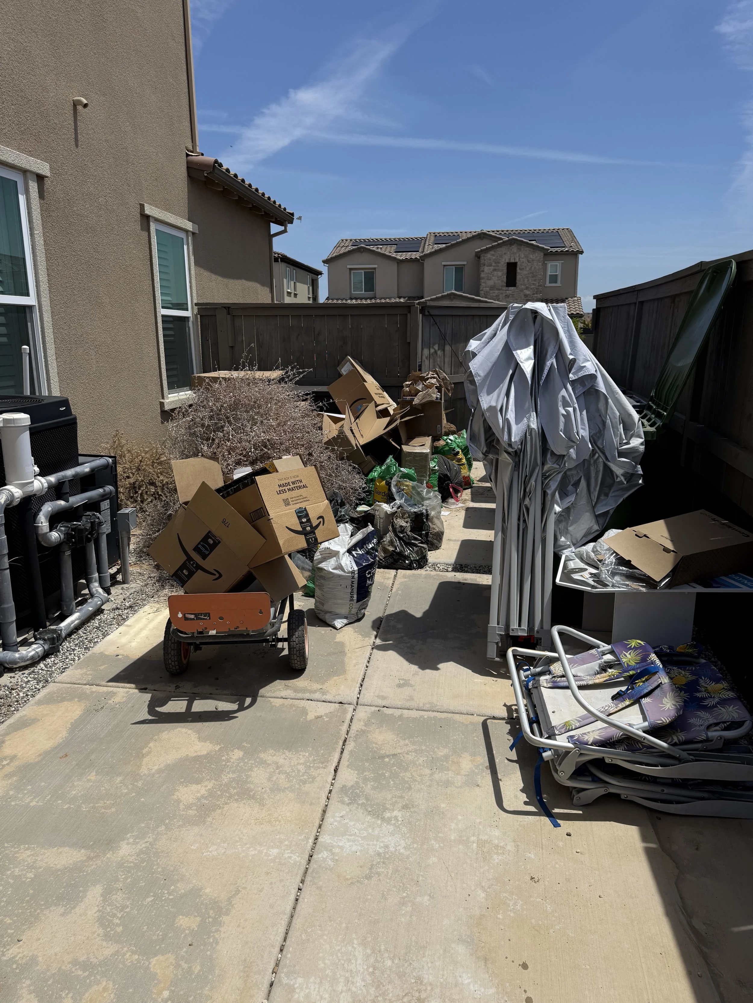 A cluttered backyard with cardboard boxes, bags, a clothes drying rack with clothes, a small orange cart, a black plastic container, and various household items. There is a wooden fence, neighboring houses with solar panels, and a clear blue sky.