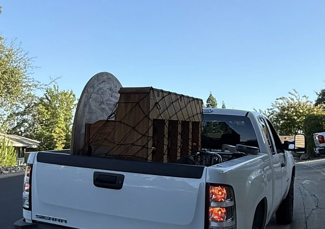 White pickup truck with loaded furniture, including a wooden chair and a large rocking chair, secured with a net, parked on a street with trees and clear blue sky