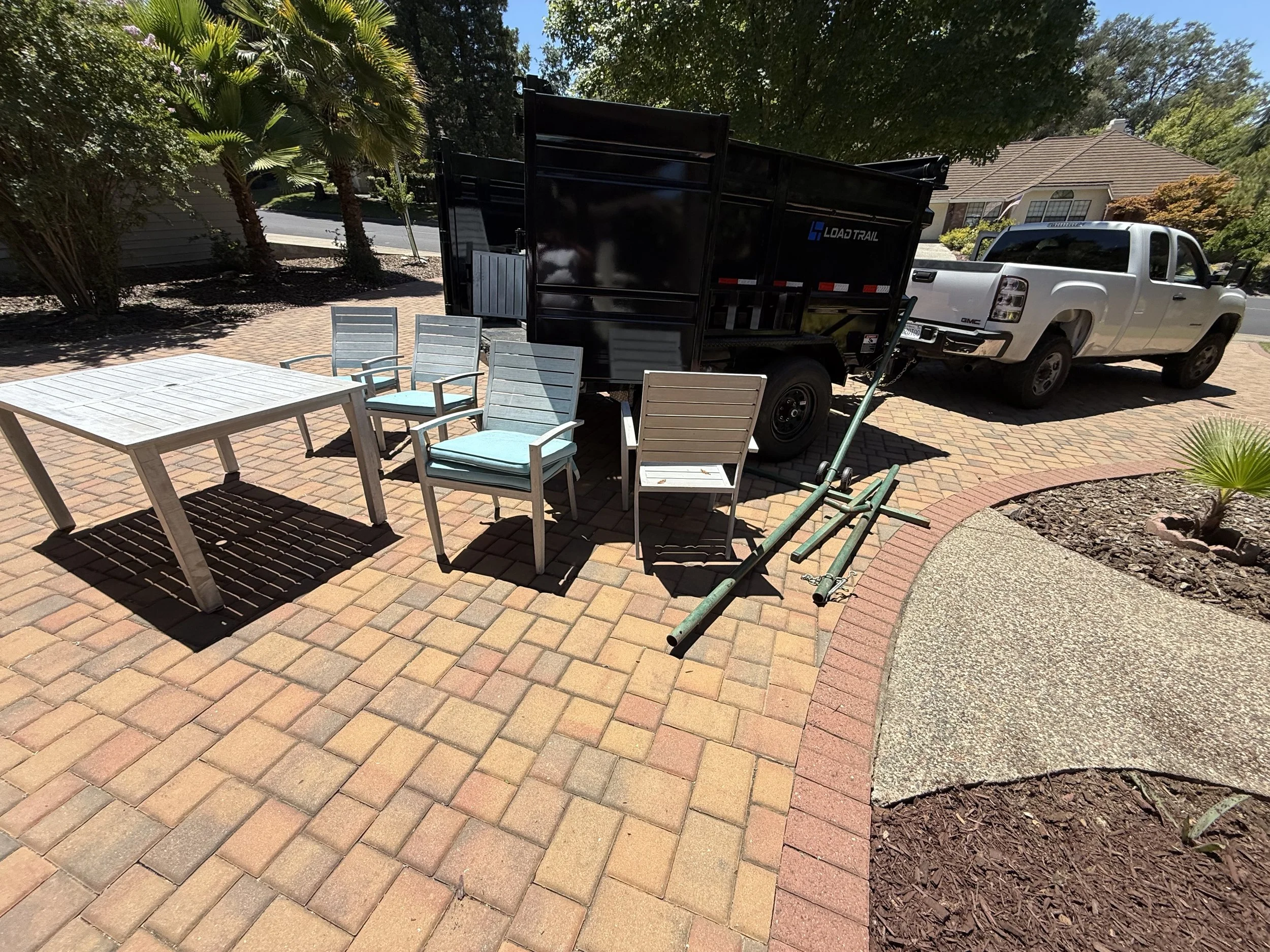 A pickup truck with a black dump trailer attached parked on a brick driveway next to a house with a garden. The scene includes outdoor furniture and lush green trees and bushes.