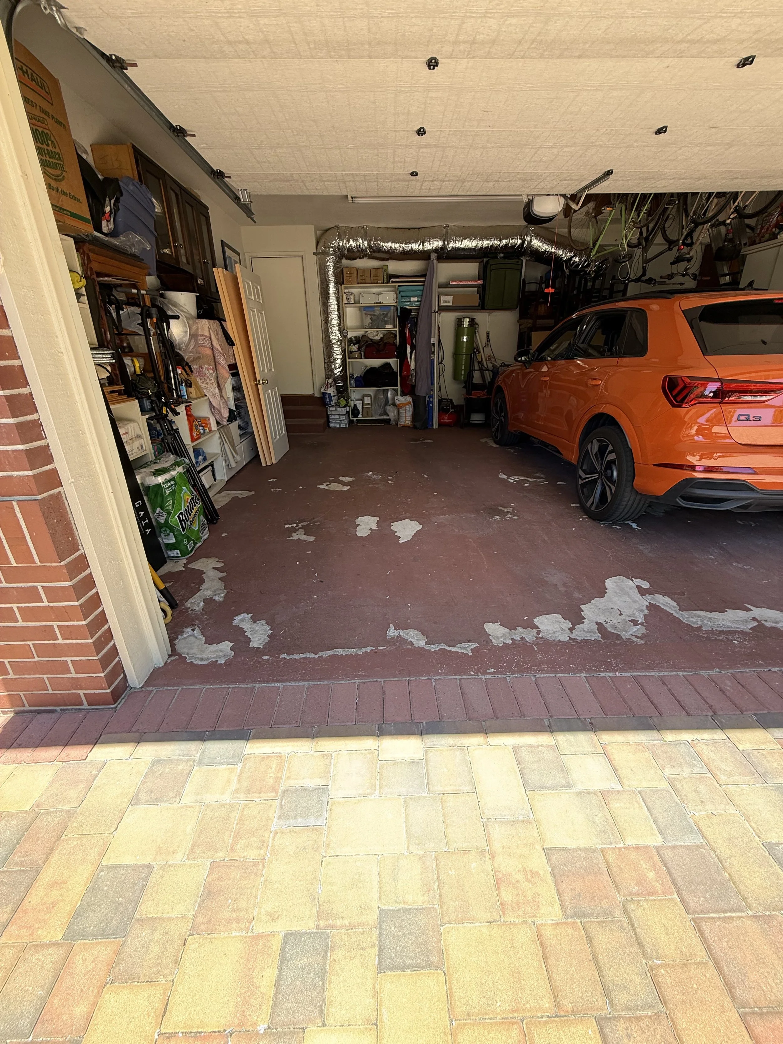 View of a garage with an orange car parked inside, various tools and storage shelves, some bicycles hanging from the ceiling, and a brick and tile driveway outside.