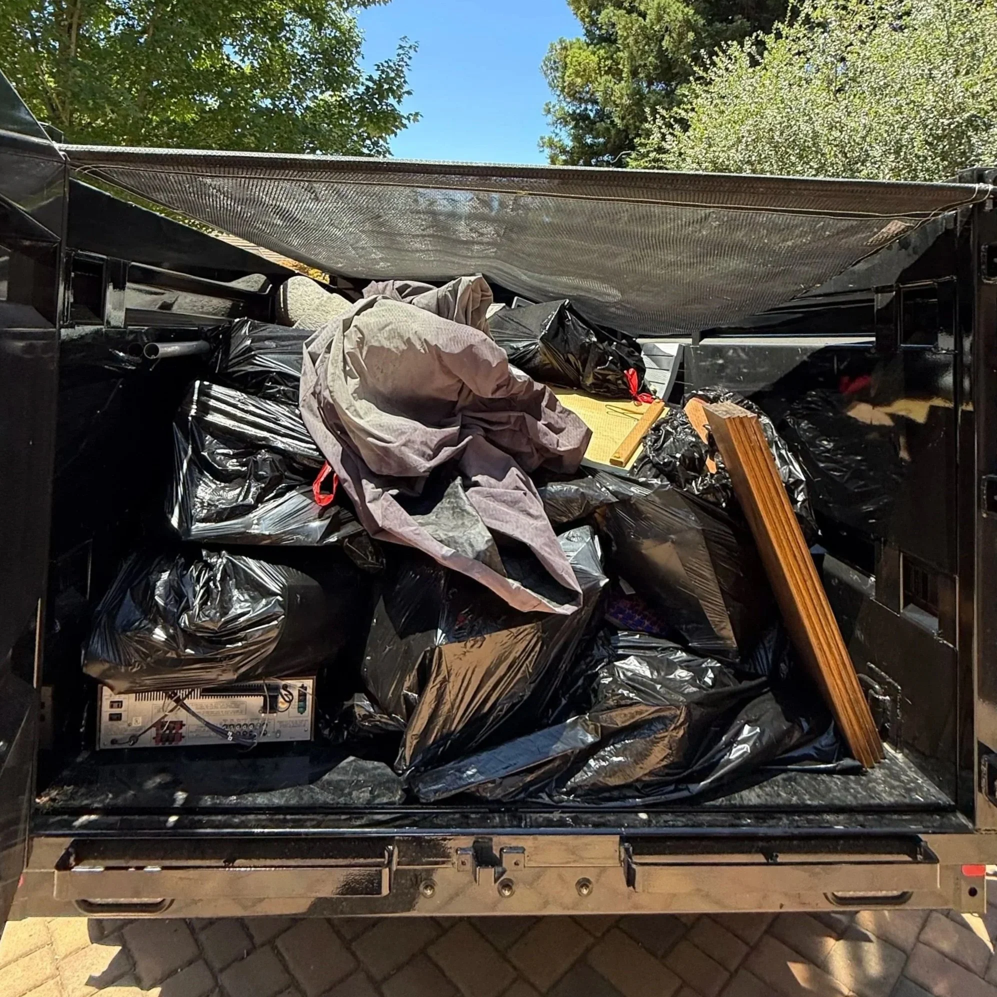 Electrical components and wires inside an electrical cabinet or panel, covered with a black plastic bag and aluminum foil, with construction materials and tools nearby. Trees and blue sky are visible outside.