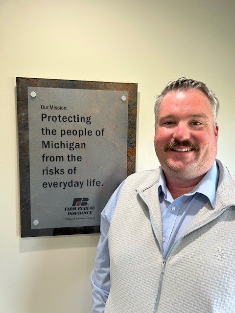 A man with a beard and mustache wearing a light blue shirt and a white vest smiling next to a mission statement sign on the wall.
