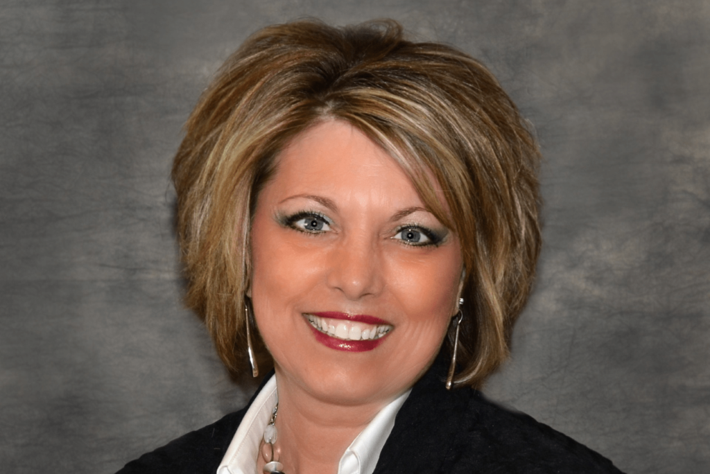 Portrait of a woman with shoulder-length light brown hair, wearing a black blazer, white shirt, and silver jewelry, smiling against a gray background.