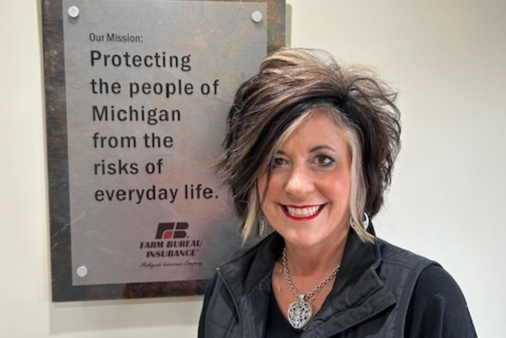 A smiling woman with short, spiky hair and a necklace stands in front of a sign that says Farm Bureau Insurance, with the mission statement about protecting Michigan residents from everyday risks.