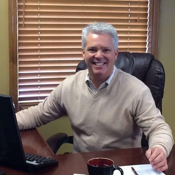 A smiling man with gray hair sitting at a desk in an office with wooden blinds behind him.