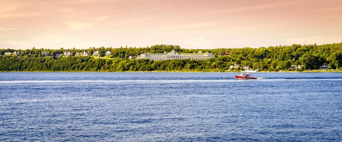 A scenic view of a large body of water with a boat and a shoreline with houses and greenery in the background during sunset.