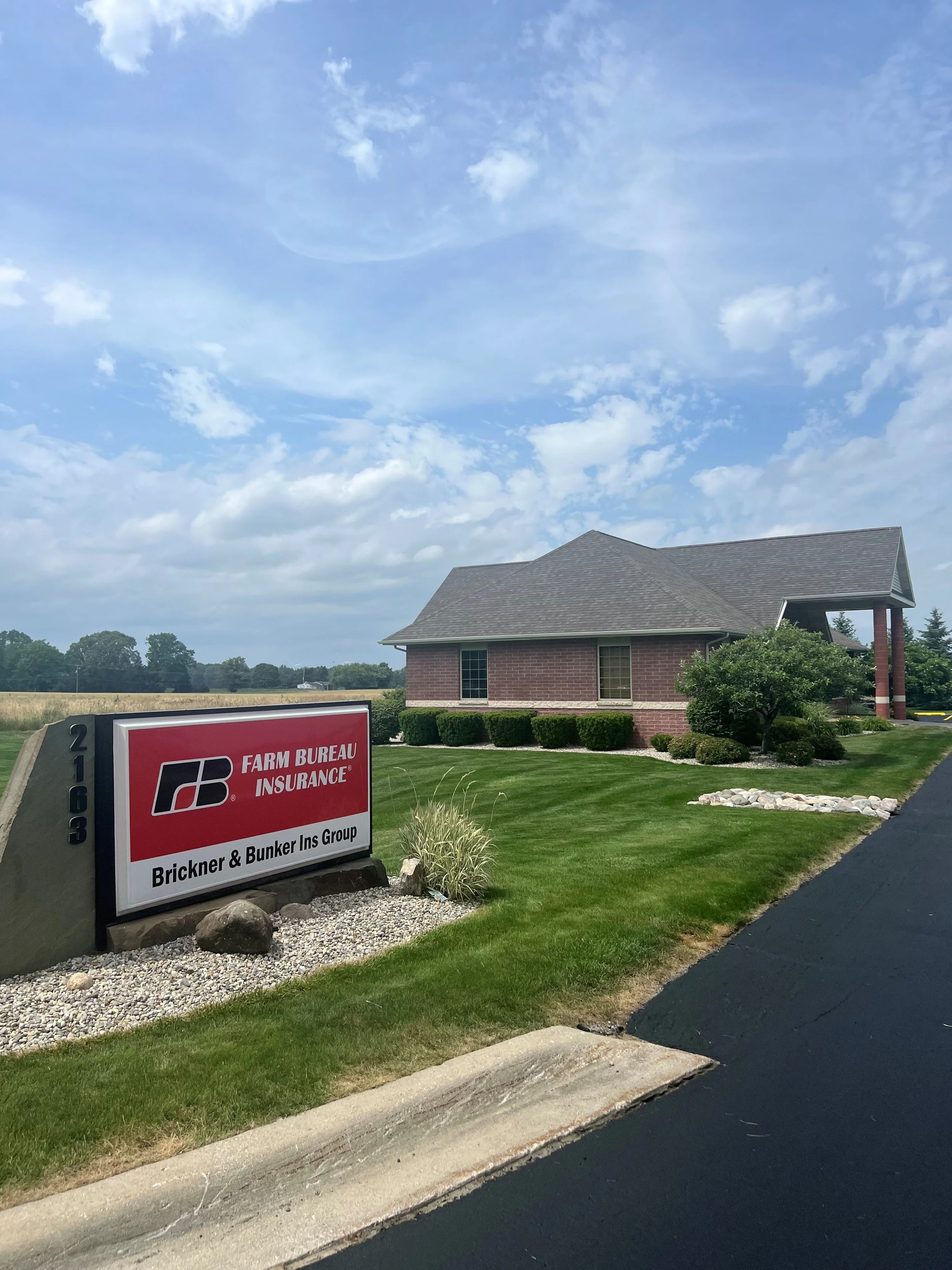 A sign for Farm Bureau Insurance in front of a brick building with a peaked roof, surrounded by neatly trimmed bushes and a well-maintained lawn. The sky is partly cloudy.