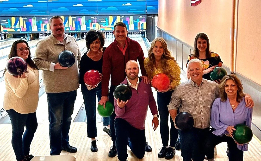 Group of ten people at a bowling alley, holding colorful bowling balls, smiling for the camera.