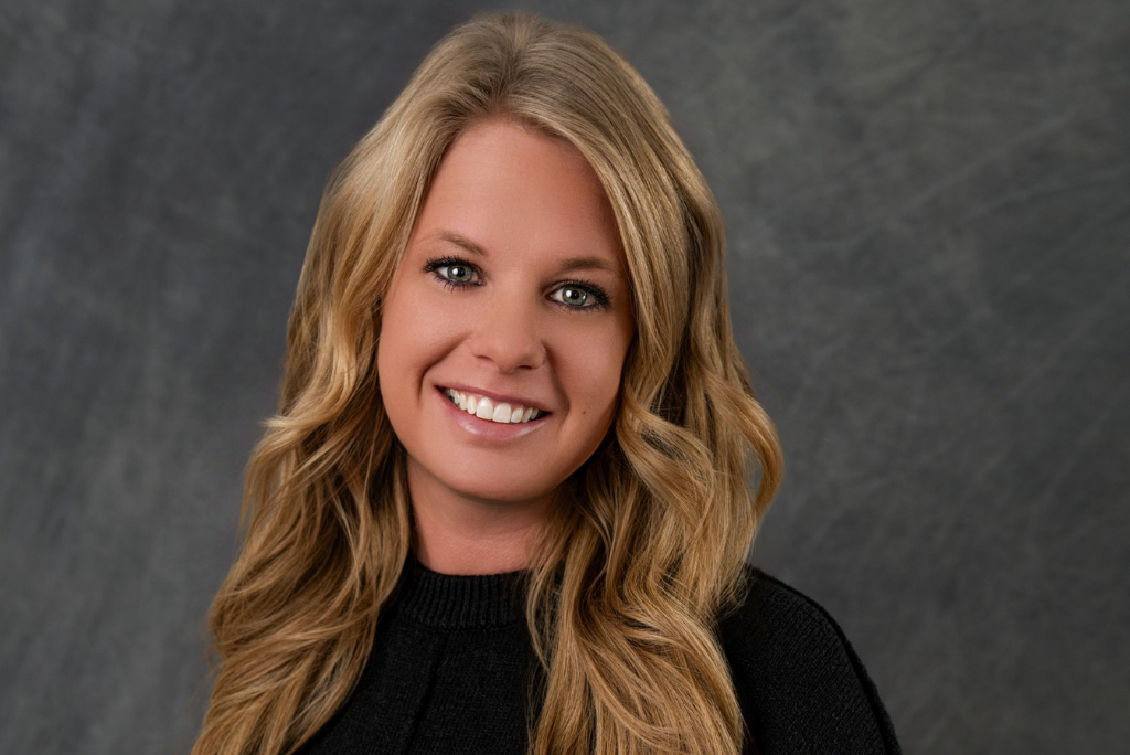 Headshot of a woman with long wavy blonde hair, smiling, wearing a black top, against a dark gray background.