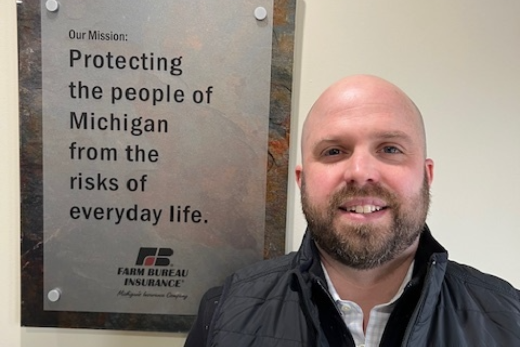 A man with a bald head and beard smiling in front of a sign that reads 'Our Mission: Protecting the people of Michigan from the risks of everyday life.' The sign also has the Farm Bureau Insurance logo at the bottom.