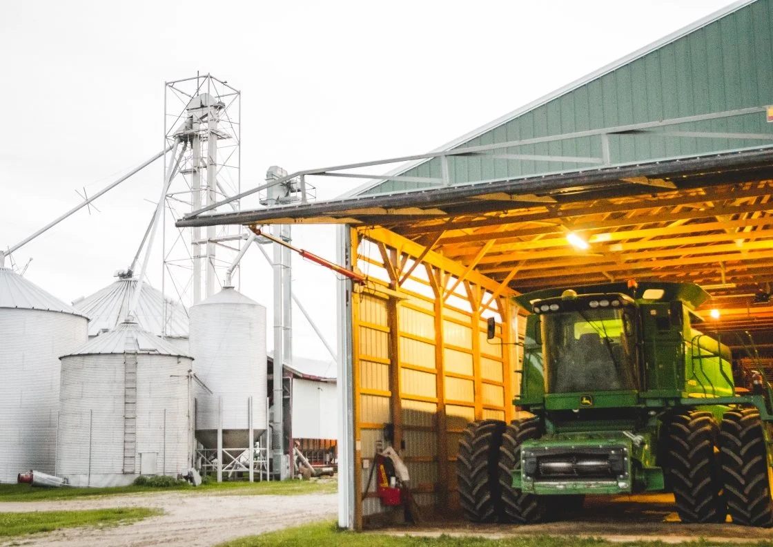 Green tractor parked inside a large wooden shed on a farm, with silos and grain storage structures in the background.