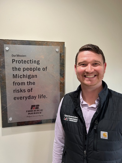 Man smiling next to a sign that states the mission of protecting Michigan residents from everyday life risks, with the Farm Bureau Insurance logo.