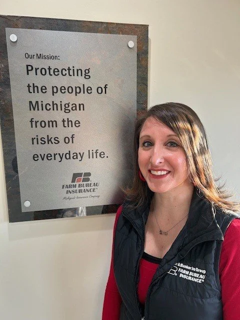 woman smiling standing next to a sign that reads Mission: Protecting the people of Michigan from the risks of everyday life. Farm Bureau Insurance logo.