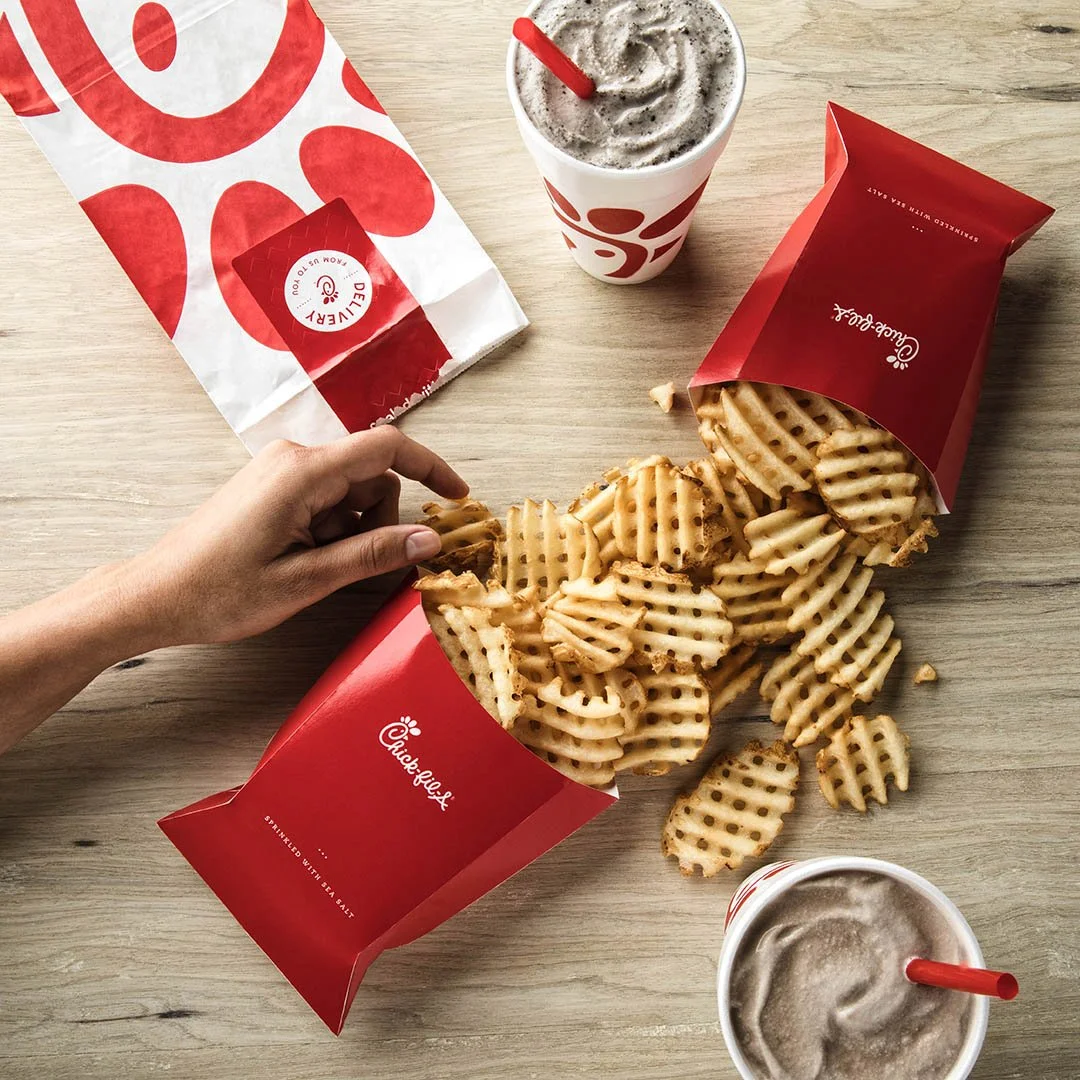 A hand reaches for waffle cut fries spilling out of a red Chick-fil-A fries container on a wooden table. Two Chick-fil-A drinks with foam and straws and a Chick-fil-A paper bag are also on the table.