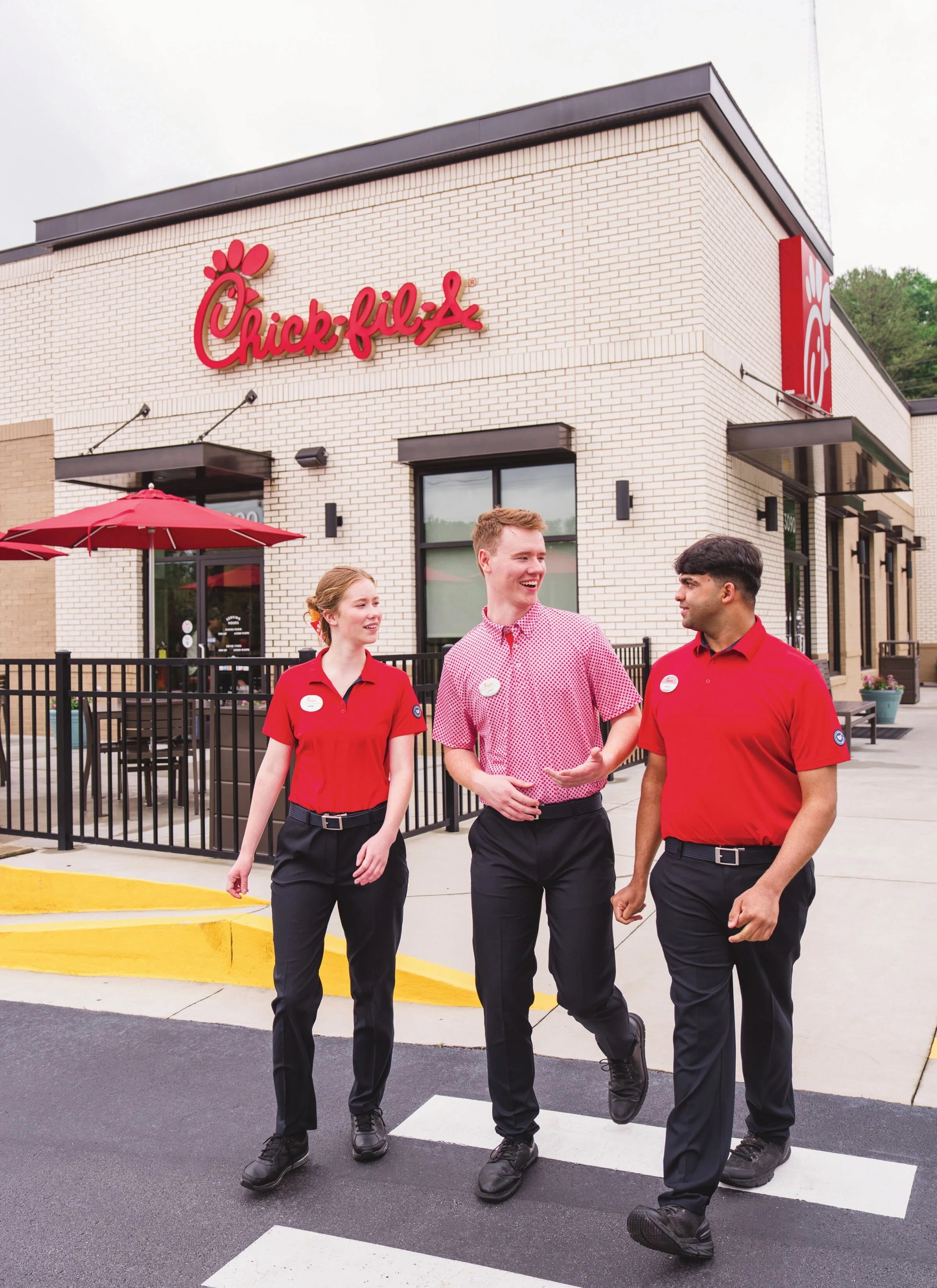 Five Chick-fil-A employees walking and talking outside a restaurant, some smiling, wearing red shirts and nametags.